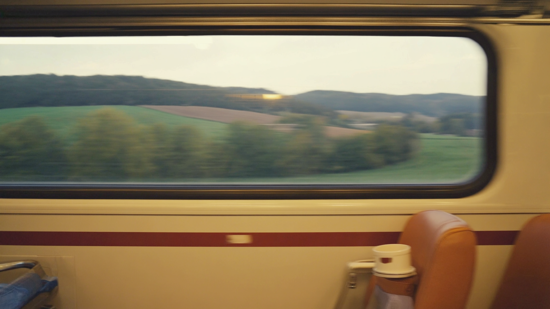 Zugfenster mit Bewegungsunschärfe, in dem sich das warme Wageninnere geisterhaft über die abendliche Landschaft legt